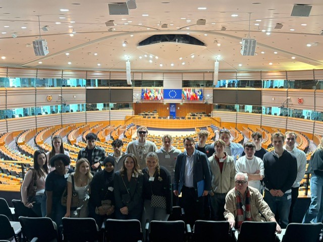 Grupp elever och vuxna står i Europaparlamentets plenisal framför halvcirkelformade sittplatser, med EU‑flagga och medlemsländernas flaggor i bakgrunden.