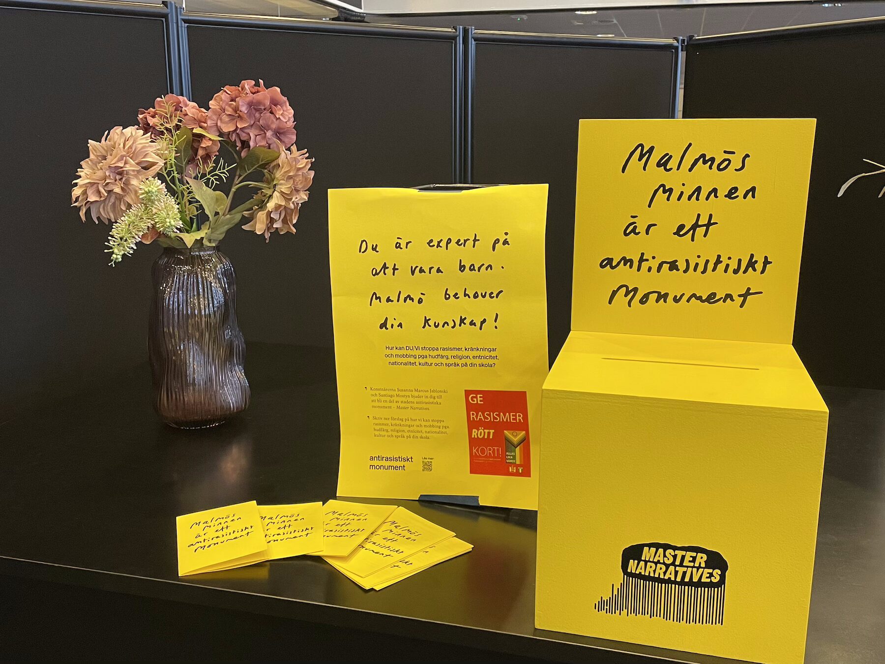Informationsmaterial om Malmös minnen är ett antirasistiskt monument med gula insamlingslådor, informationsblad och kort på ett bord bredvid en blomvas.