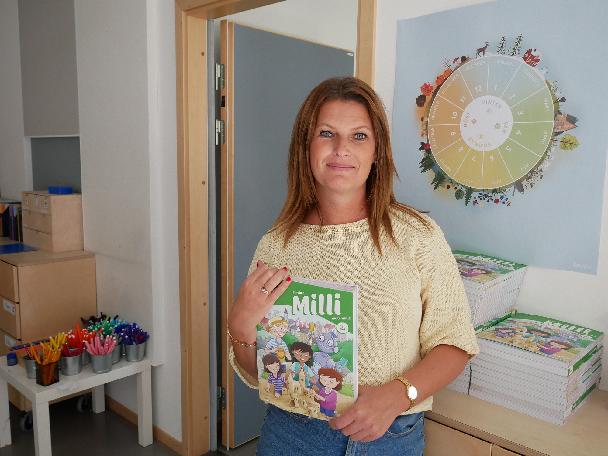 En kvinna står i ett klassrum och håller i en mattebok för lågstadieelever.