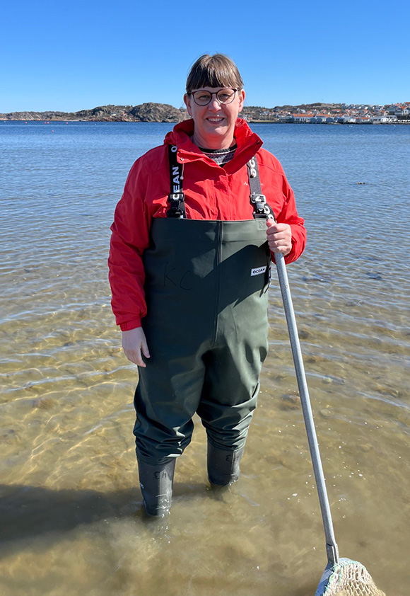 En kvinna i röd jacka och i vadarbyxor står i vatten med en hov.