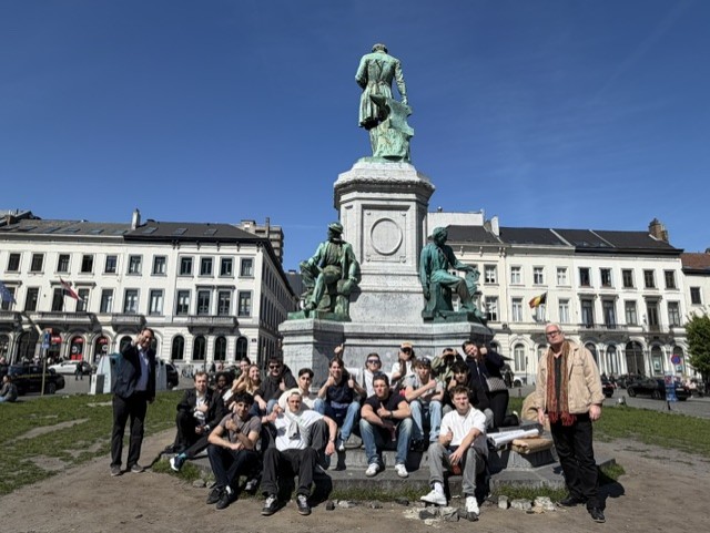 Grupp personer sitter och står framför ett monument på ett torg omgivet av byggnader under klarblå himmel.