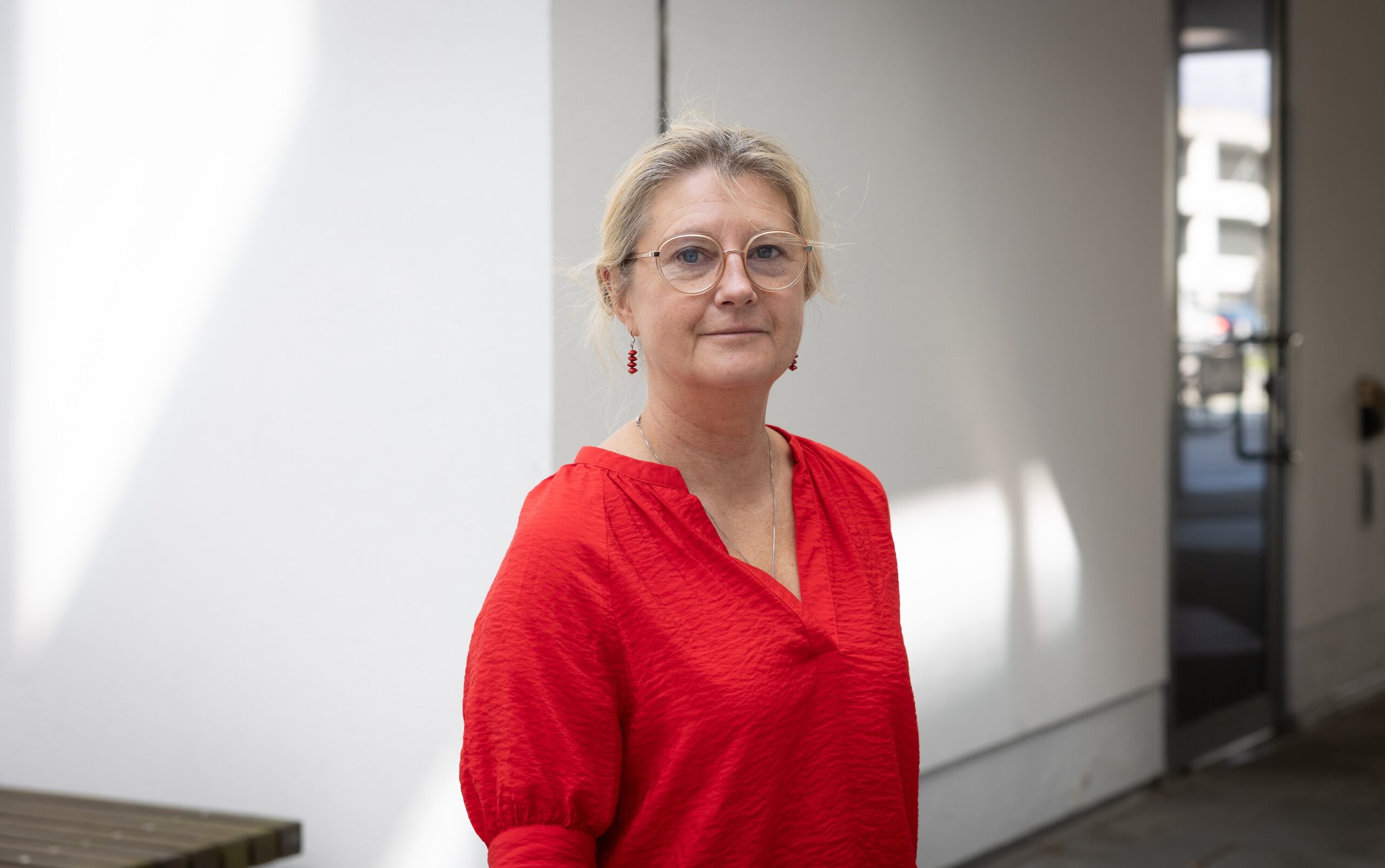 Person i röd blus och glasögon står i en ljus inomhusmiljö med vita väggar och glasdörr i bakgrunden.