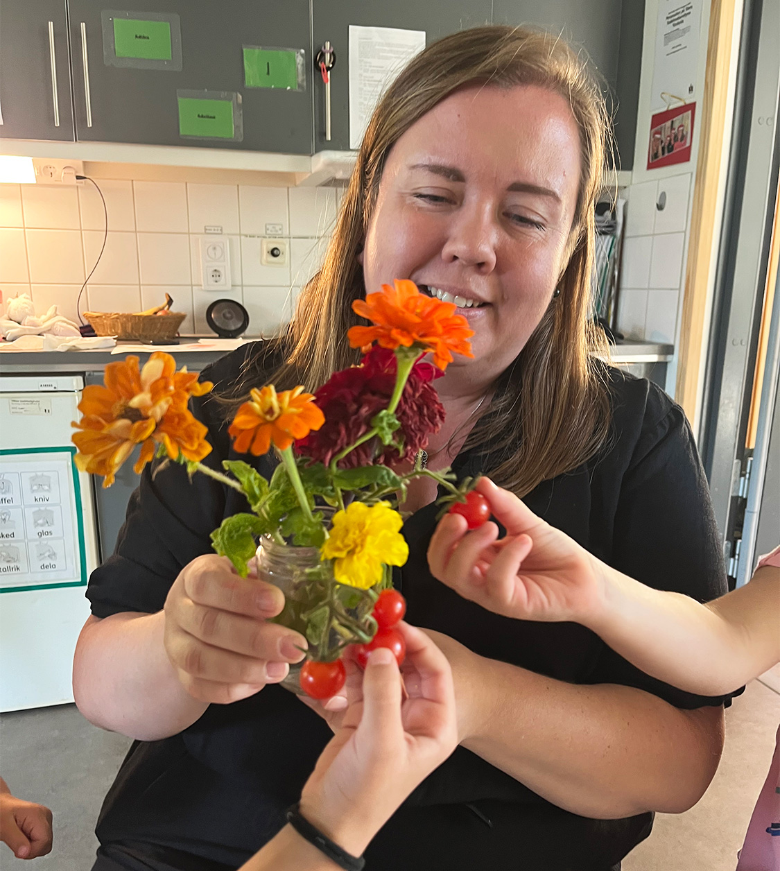 En kvinnlig pedagog sitter med en vas med oranga och gula blommor. I vasen finns även en tomatkvist. Några barnhänder plockar tomaterna. Kvinnan ler varmt.