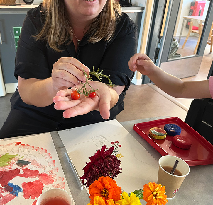 En kvinnlig pedagog sitter vid ett bord och håller en tomatkvist i sin hand. En barnhand sträcker sig mot tomaten.