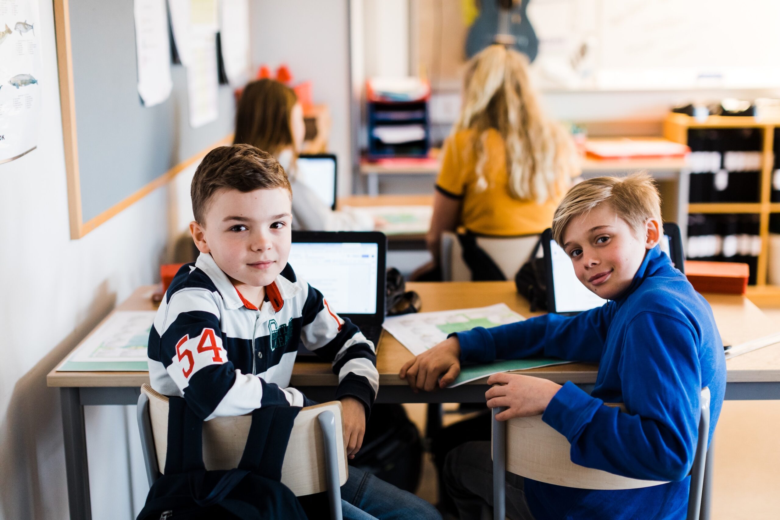 Elever arbetar vid datorer i ett klassrum. Två elever sitter vid ett bord i förgrunden och har bärbara datorer och arbetsmaterial framför sig, medan fler elever arbetar vid sina platser i bakgrunden.