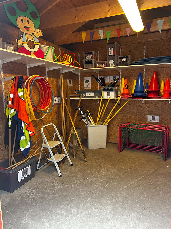 Insidan av en lekbod med organiserad rastutrustning. På hyllor längs väggarna finns rockringar, koner, racketar, hopprep och förvaringslådor. En stor figur av Super Mario-svampen pryder en hylla. På golvet står små mål, käppar och annat material för lekar och aktiviteter. Utrustningen är uppmärkt och sorterad för enkel utlåning under raster.